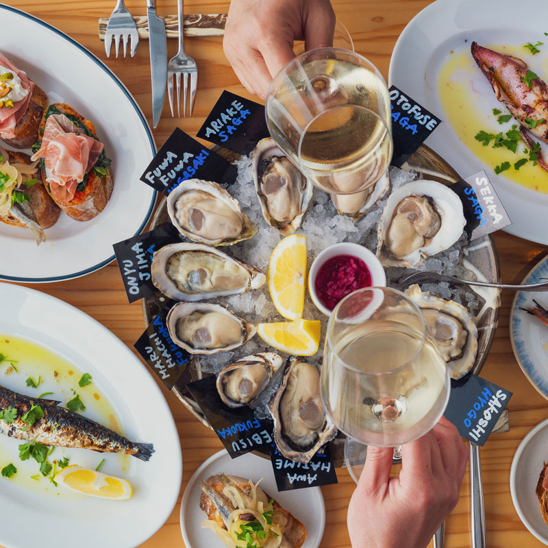 BACARO CARBON oyster barのKUOHKA店舗ページに掲載された料理写真：殻付き牡蠣の盛り合わせとグラス、小皿料理